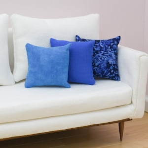May include: A white couch with three blue throw pillows. The couch has a white, textured fabric and wooden legs. The pillows are different shades of blue and have different patterns.