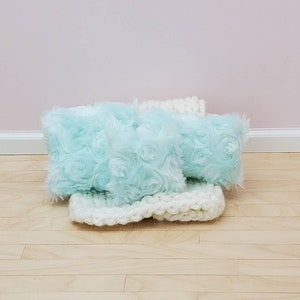 May include: Three light blue fluffy pillows with a white crocheted blanket. The pillows have a rosette pattern.