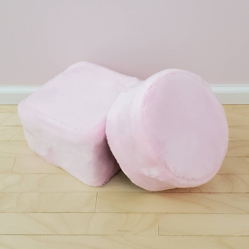 K&ouml;nnte beinhalten: Zwei hellrosa Poufs. Einer ist quadratisch, der andere zylindrisch. Beide bestehen aus einem weichen, flauschigen Material. Die Poufs stehen vor einer hellrosa Wand und einem hellen Holzboden.