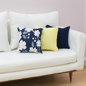 May include: A white sofa with three decorative throw pillows. One pillow is navy blue with a white floral pattern, a second is yellow, and the third is solid navy blue. The sofa has wooden legs.