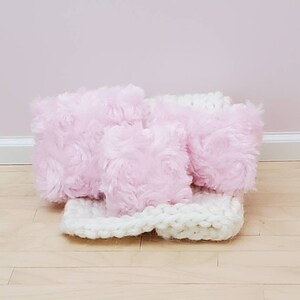May include: Three pink fluffy cushions with a white knitted throw. The cushions have a rose-like texture.