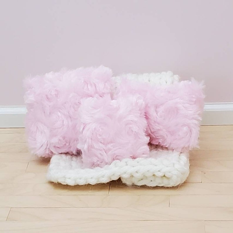 May include: Three pink fluffy cushions with a white knitted throw. The cushions have a rose-like texture.