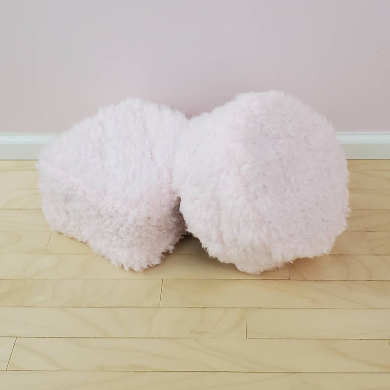 K&ouml;nnte beinhalten: Zwei flauschige, hellrosa Poufs. Die Poufs bestehen aus einem weichen, strukturierten Material und stehen auf einem hellen Holzboden. Der Hintergrund ist eine einfarbige hellrosa Wand.