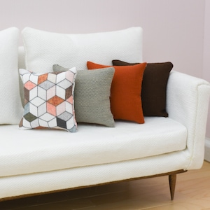 May include: A white couch with four throw pillows. The pillows are patterned with geometric shapes in shades of orange, brown, and gray.