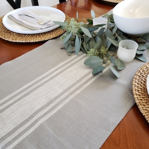 May include: A gray table runner with white stripes, a wooden table, and a centerpiece of eucalyptus leaves and white candle holders.