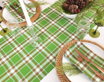 May include: A green and white checked table runner with red accents. The runner is laid on a white tablecloth and features a white candle holder with a white candle. There are two place settings with white plates, woven placemats, green glasses, and cutlery.