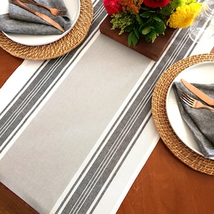 May include: A table setting with a white and gray striped table runner, two woven placemats, white plates, gray napkins, and copper silverware. A bouquet of orange, red, and yellow flowers is in the center of the table.