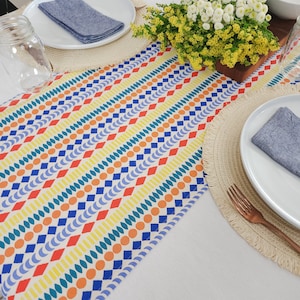 May include: A white tablecloth with a colorful geometric patterned table runner. The runner has blue, orange, yellow, and green geometric shapes. There are two place settings with white plates, blue napkins, and woven placemats.