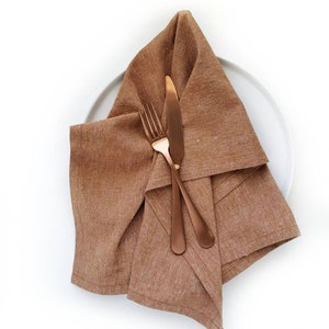 A caramel colored linen towel is shown on a white plate with coppery-colored silverware.