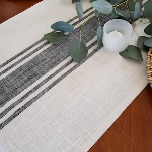 May include: A white linen table runner with black stripes. The runner is on a wooden table with a small white candle holder and eucalyptus leaves.