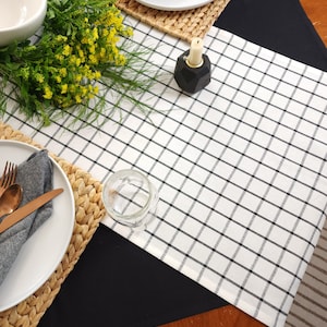 May include: A white table runner with black checkered pattern, placed on a black tablecloth. The runner is set with a white plate, a gray napkin, and gold silverware. A glass jar and a black candle holder are also on the table.