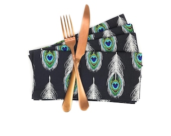May include: A set of black napkins with a peacock feather pattern, folded and placed on a white surface. A gold fork and knife are placed on top of the napkins.