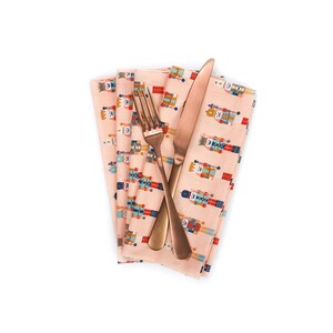 May include: A pink fabric napkin with a pattern of nutcracker soldiers. A gold fork and knife are placed on top of the napkin.