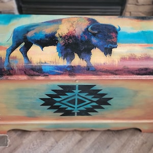 May include: A wooden storage chest with a hand-painted design of a blue and orange buffalo on a yellow and blue background. The chest also features a black and turquoise geometric pattern on the front.