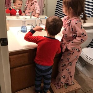 Puede incluir: Dos niños cepillándose los dientes en un baño. Uno lleva una camisa roja y pantalones a rayas azules, de pie sobre un taburete de madera. El otro niño lleva un pijama rosa con estampado. Un espejo refleja la escena.