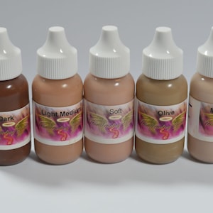 May include: Five small bottles of liquid foundation in various shades: Dark, Light Medium, Soft, Olive, and Medium Fair. Each bottle has a white cap and a label with the words "Transcending Beauty" and the shade name.
