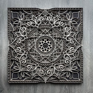 May include: A black and white intricate mandala design carved into a wooden panel. The design features a central circle with radiating patterns and a square frame.