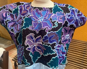 May include: A short-sleeved blouse featuring a black base with an intricate, multi-colored floral pattern in shades of purple, blue, and teal. The garment has a square neckline and a solid light blue fabric panel attached at the bottom hem.