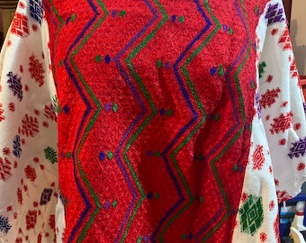 May include: A white blouse featuring a large red central panel with a vibrant zigzag pattern in green and blue. The sleeves and sides are white with scattered, small geometric embroidery in red, green, blue, and purple throughout the fabric.