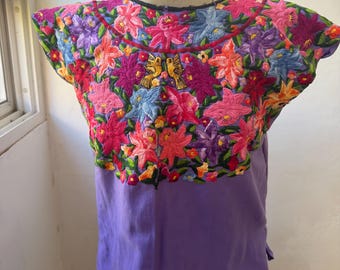 May include: A short-sleeved lavender blouse featuring an intricate, multi-colored floral embroidery pattern across the chest and shoulders. The design includes vibrant pink, purple, and blue flowers, along with two small yellow birds centered.