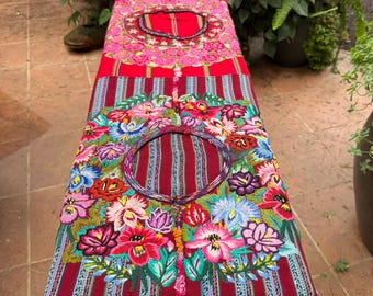 May include: A long, rectangular textile runner featuring a deep red base with vertical stripes and three circular openings. The fabric is decorated with vibrant, multi-colored floral embroidery in shades of pink, blue, and green, finished with fringe.