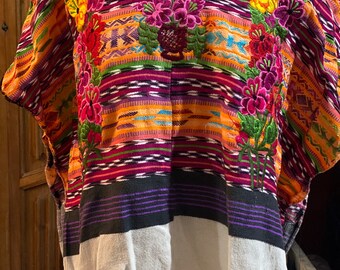 May include: A colorful woven blouse featuring intricate floral embroidery in shades of pink, red, yellow, and green. The garment displays horizontal stripes in orange, purple, and black, transitioning into a solid white panel at the lower section.