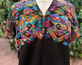May include: A black blouse featuring a V-neckline and short sleeves, adorned with intricate, multi-colored geometric embroidery across the chest and shoulders. The embroidery incorporates vibrant shades of red, blue, yellow, and green thread.