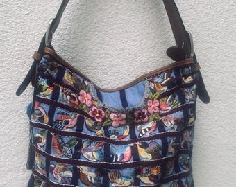Pode incluir: Uma bolsa de ombro estilo hobo com corpo de tecido às riscas azuis e pretas com bordados coloridos de pássaros e flores. A bolsa inclui uma base de couro castanho sólido, alças de couro castanho com ferragens prateadas e uma borla azul.