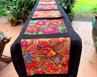 May include: A black table runner with vibrant floral and bird embroidery. The runner features a repeating pattern of colorful flowers and birds in shades of red, pink, orange, and green. The edges are trimmed with a black border.