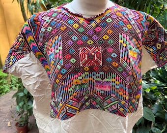 May include: A colorful, embroidered blouse with a v-neckline. The top portion features a vibrant geometric pattern in shades of red, purple, yellow, green, and blue. The bottom is a plain, off-white fabric. The blouse is displayed on a mannequin.