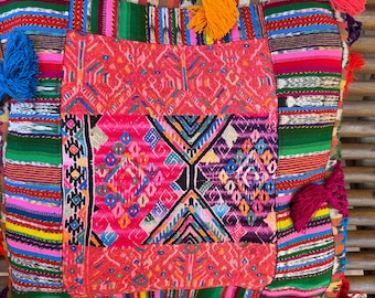 May include: A square, vibrantly colored pillow with a patchwork design. The pillow features a central geometric pattern in pink, purple, and blue, surrounded by striped and patterned borders. Colorful tassels adorn the edges, adding a decorative touch.