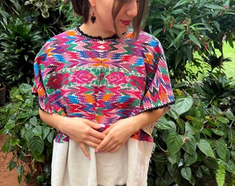 May include: A short-sleeved blouse featuring a vibrant, multi-colored woven pattern with zig-zag and floral motifs in shades of pink, blue, orange, and green, finished with a black scalloped trim along the neckline and sleeve edges.