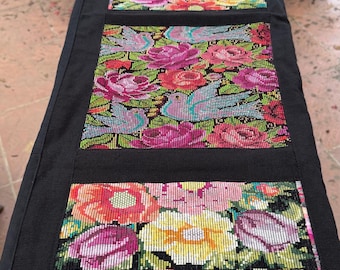 May include: A rectangular table runner with a floral design. The runner features a black border and three panels of colorful flowers in shades of red, pink, yellow, and purple. The flowers are densely packed and create a vibrant pattern.