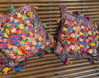 May include: Two colorful, square decorative pillows with vibrant geometric patterns in pink, yellow, blue, and orange. Each pillow has a fringe of tassels in various colors. The pillows are placed on a wooden slatted surface.