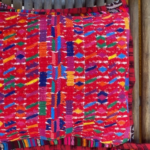 May include: A square cushion featuring a red background and a vibrant, geometric pattern. The design incorporates various shapes and colours, including blue, yellow, green, and purple. The cushion is placed on a wooden bench.