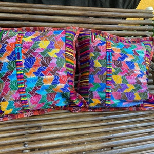 Puede incluir: Dos cojines decorativos coloridos y tejidos a mano con patrones geométricos y flecos. Los cojines están hechos de una tela suave y texturizada y presentan una mezcla vibrante de colores, incluyendo rojo, azul, verde, amarillo y morado.