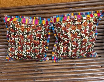 May include: Two square throw pillows with a colorful bird and floral pattern. The pillows have a white background with brown birds and orange flowers. The edges are trimmed with a vibrant, striped fabric.