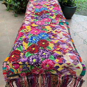 May include: A colorful table runner with a floral pattern and fringe. The runner is made of woven fabric and features a variety of colors, including red, pink, purple, blue, yellow, and green.