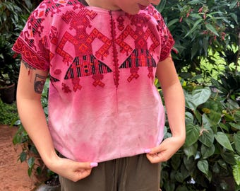 May include: A short-sleeved pink blouse featuring intricate red and black geometric embroidery across the chest and shoulders. The garment has a round neckline and a central vertical seam, with a subtle ombre effect fading from dark pink to light pink.