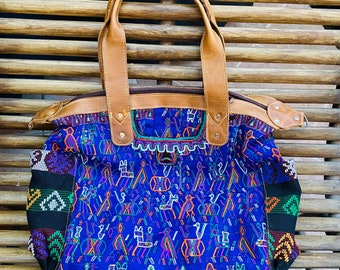 May include: A blue tote bag with a colorful woven pattern featuring figures and animals. The bag has brown leather handles, trim, and a zipper closure. The bag is sitting on a wooden bench.