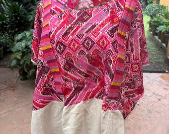 May include: A vibrant pink and red huipil tunic featuring intricate geometric patterns and a floral embroidered neckline. The garment includes wide, striped shoulder panels and a solid off-white fabric section at the bottom hem, displayed on a mannequin.