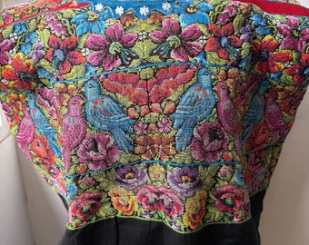 May include: A short-sleeved blouse featuring intricate, multi-colored embroidery of birds and floral patterns. The design includes vibrant shades of red, blue, purple, and yellow on a black base, with a blue neckline accented by small white flowers.