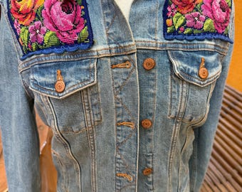 May include: A light blue denim jacket featuring two rectangular patches on the chest area. Each patch displays a vibrant floral needlepoint design with pink, orange, and yellow roses, accented by green leaves and a blue scalloped border trim.
