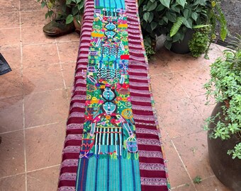 May include: A long, rectangular textile runner featuring a central panel with intricate, colorful embroidery depicting stylized figures and geometric patterns, flanked by deep maroon and white striped fabric borders with fringed ends.
