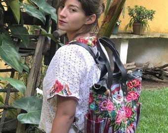 May include: A backpack featuring a black leather top and straps, with a lower section made of burgundy and white striped fabric. The bag is decorated with vibrant, multi-colored floral embroidery and includes side pockets and black handles.
