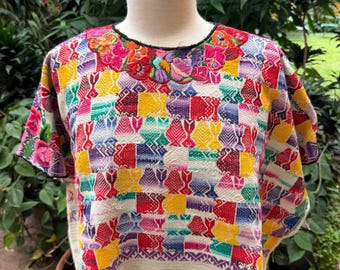 May include: A short-sleeved blouse featuring a vibrant, multi-colored geometric pattern of squares in red, yellow, blue, and purple. The neckline is adorned with detailed floral embroidery in bright hues, and the hem is a solid off-white fabric.