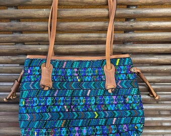 May include: A tote bag featuring a vibrant, multi-colored woven textile pattern in shades of blue, green, purple, and red. The bag includes tan leather handles and side accents with knotted leather ties on both sides, providing a structured shape.
