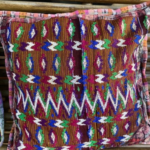 Puede incluir: Una almohada cuadrada marrón con un patrón geométrico colorido. El patrón presenta diamantes y zigzags blancos, azules, verdes y morados.