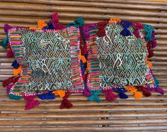 May include: Two decorative square pillows with colorful geometric patterns and tassel trim. The pillows feature a vibrant mix of colors including green, purple, orange, and blue, creating a bohemian aesthetic. The pillows are placed on a wooden surface.