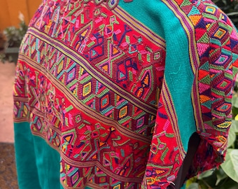 May include: A teal blouse featuring a wide, square-cut neckline and a prominent chest panel decorated with intricate, multi-colored geometric and bird-shaped embroidery in shades of red, yellow, purple, and green on a vibrant red background.